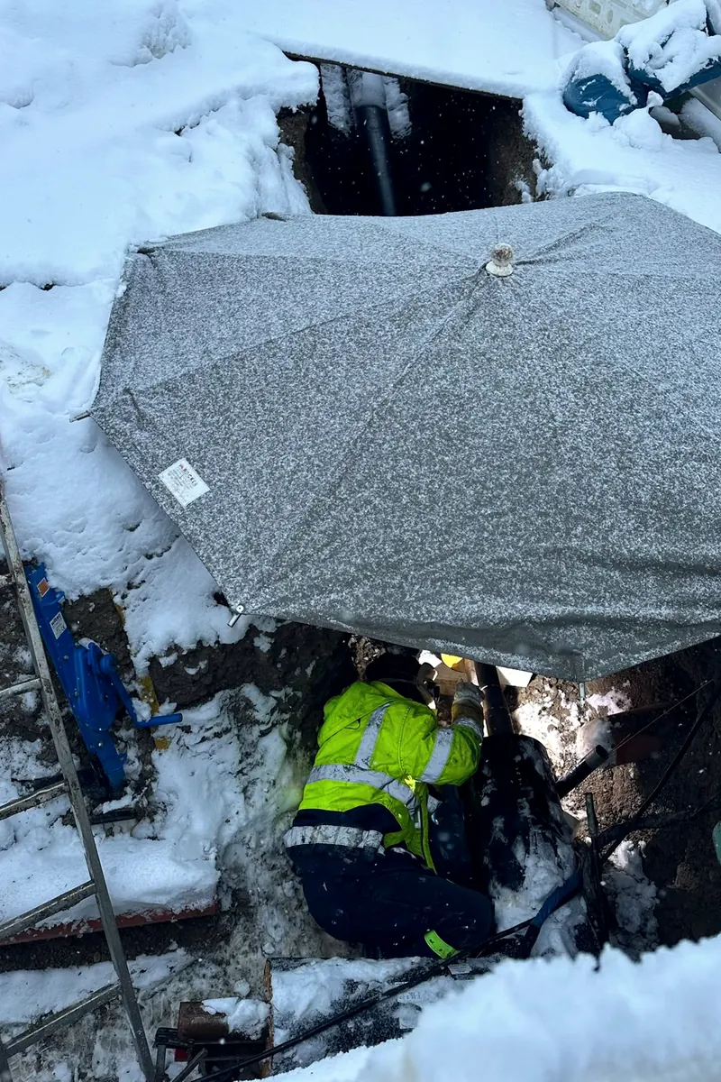 Monteur arbeitet im verschneiten Leitungsgraben unter einem Schutzschirm